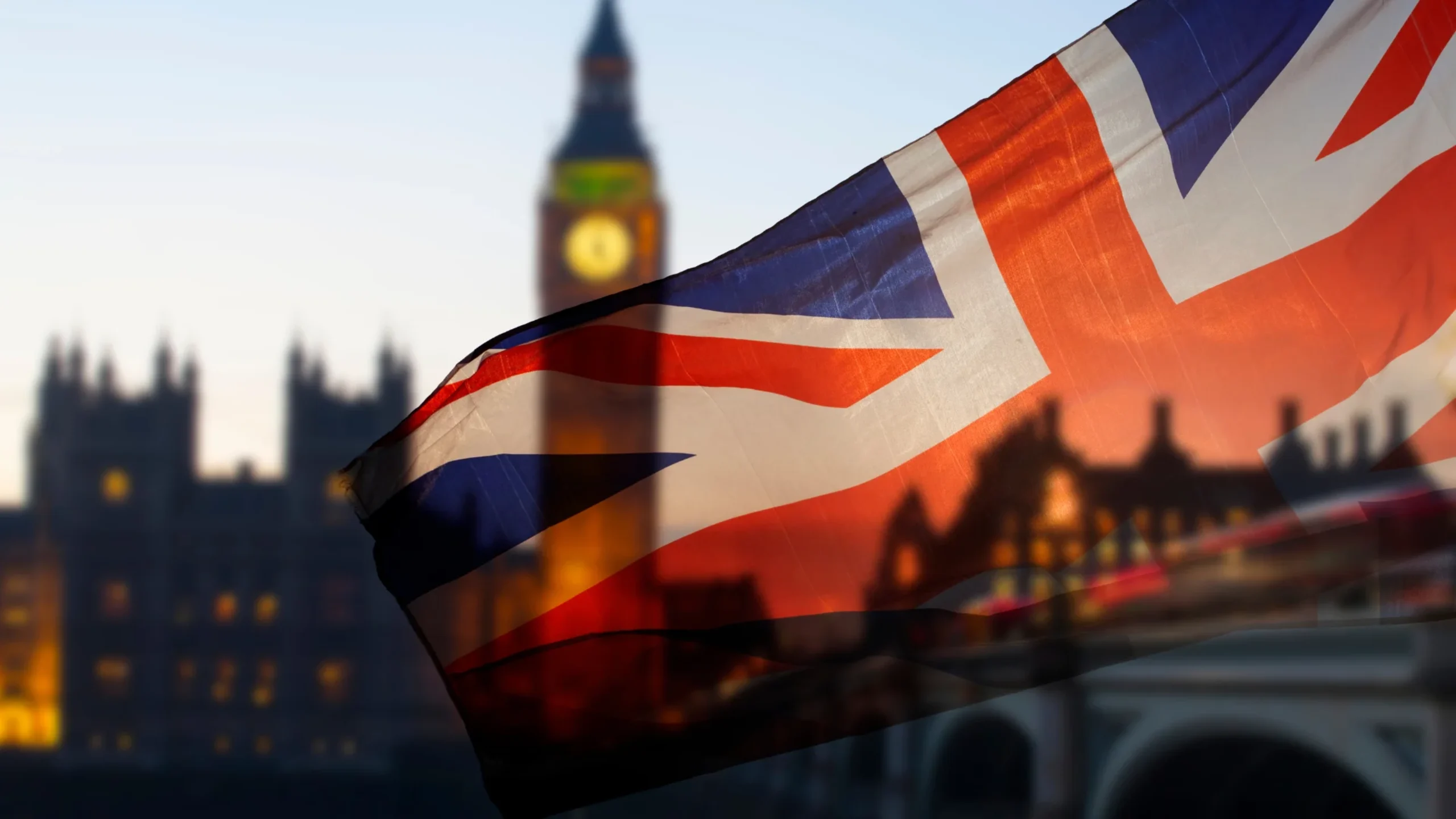 A close-up of the Union flag waving against a blurred backdrop of Big Ben and Parliament buildings at dusk