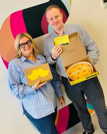Two people smiling and holding Moustache cookie boxes, one with a cookie that says “Thanks GBL,” standing in front of a colorful abstract wall design.
