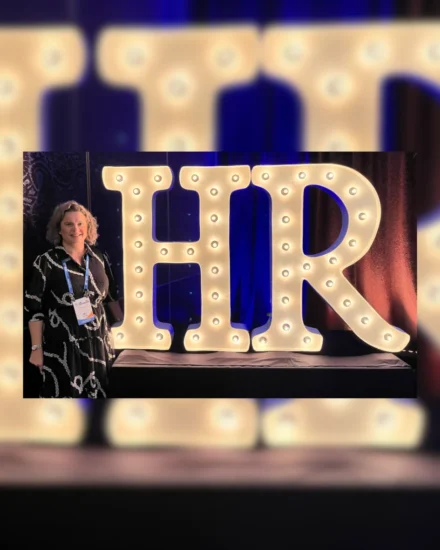 Woman standing next to large illuminated "HR" letters at an event, wearing a lanyard and smiling.