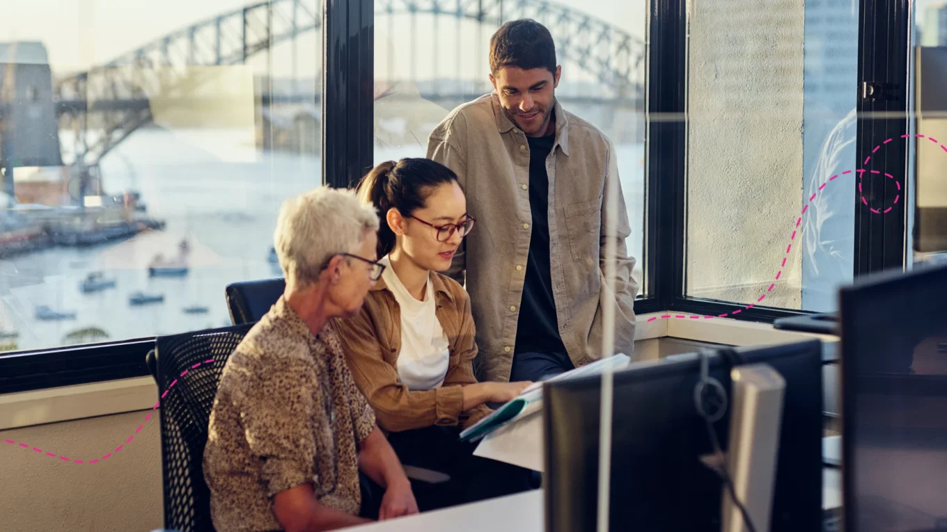 Three people looking at a computer together in an office with Sydney Harbour bridge in the background.