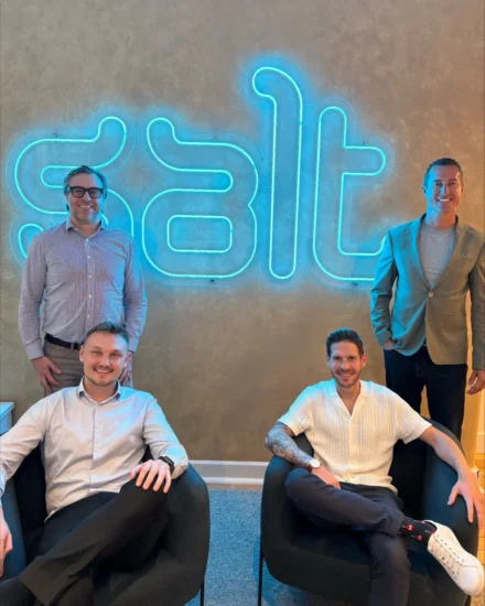 A group of four Salt employees posing in front of a glowing "salt" sign at an office, with two sitting in chairs and two standing behind them.