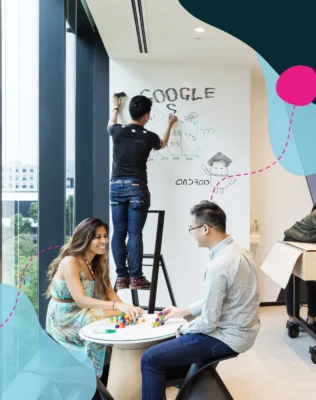 Poeple sitting at an office desk with someone else writing "Google" on a whiteboard