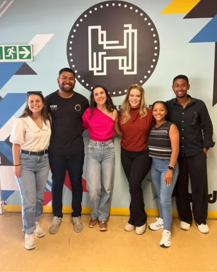 Seven smiling team members standing together in front of a wall with a large “H” logo.