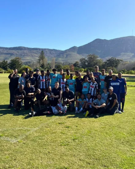 Large group of people posing on a soccer field with mountains in the background on a sunny day.