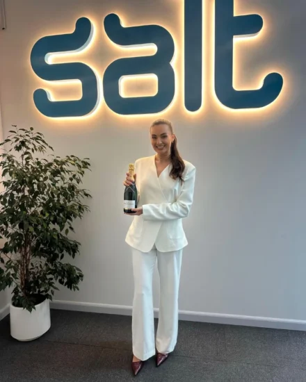 Woman in a white suit holding a champagne bottle, standing in front of an illuminated “salt” logo in an office.