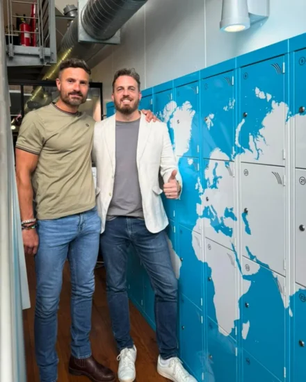 Two men, our CEO Elliot Dell and Africa Director and Founder, Alex Martin, smiling and posing indoors beside blue lockers featuring a world map design at the London office.